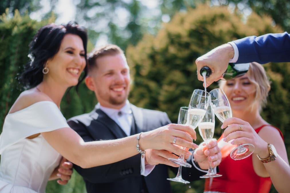 bride, groom, and woman in red dress  holding glasses and hand pouring the Champagne